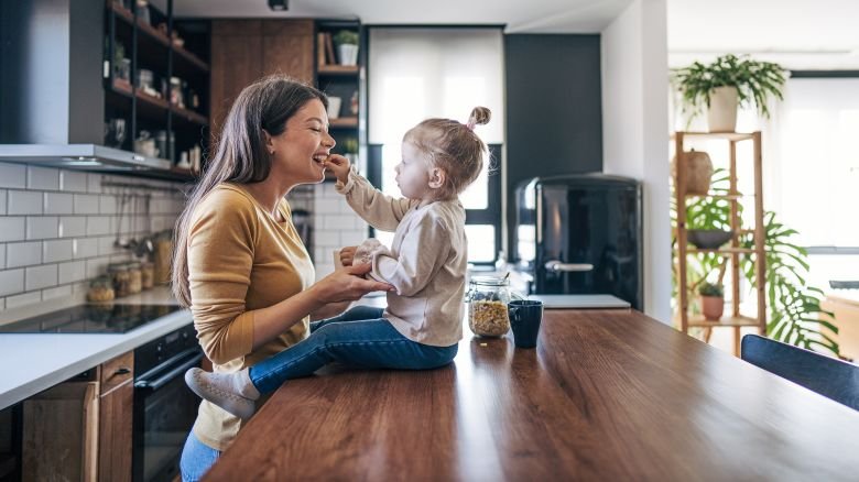 Mother and toddler sharing a joyful moment in a modern kitchen, with the child feeding the mother while both smile amidst sleek appliances and indoor plants