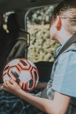 white schoolgirl in a plaid pinafore and blue uniform holds a soccer ball on her lap in the front seat as her father prepares to drive, ready for after-school sport. A candid moment linking family routine and children's sport.